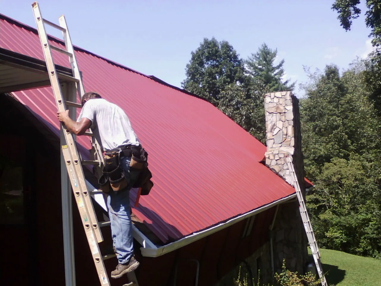 Roofer on a ladder beside a red metal roof and stone chimney — Soffit & Fascia Repair by Roofing Brothers in Tomah